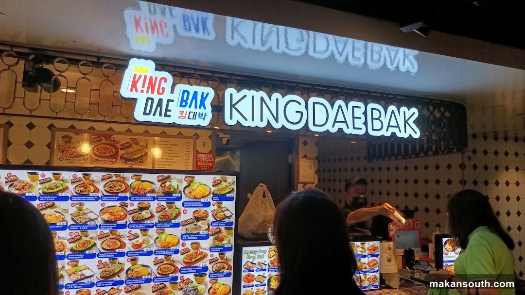 The storefront of a Korean restaurant called King Dae Bak. The restaurant has a large illuminated sign with the name of the restaurant. Below the sign is a menu board displaying various Korean dishes with pictures and prices. Several customers are visible in the foreground, looking at the menu. A staff member is seen behind the counter.