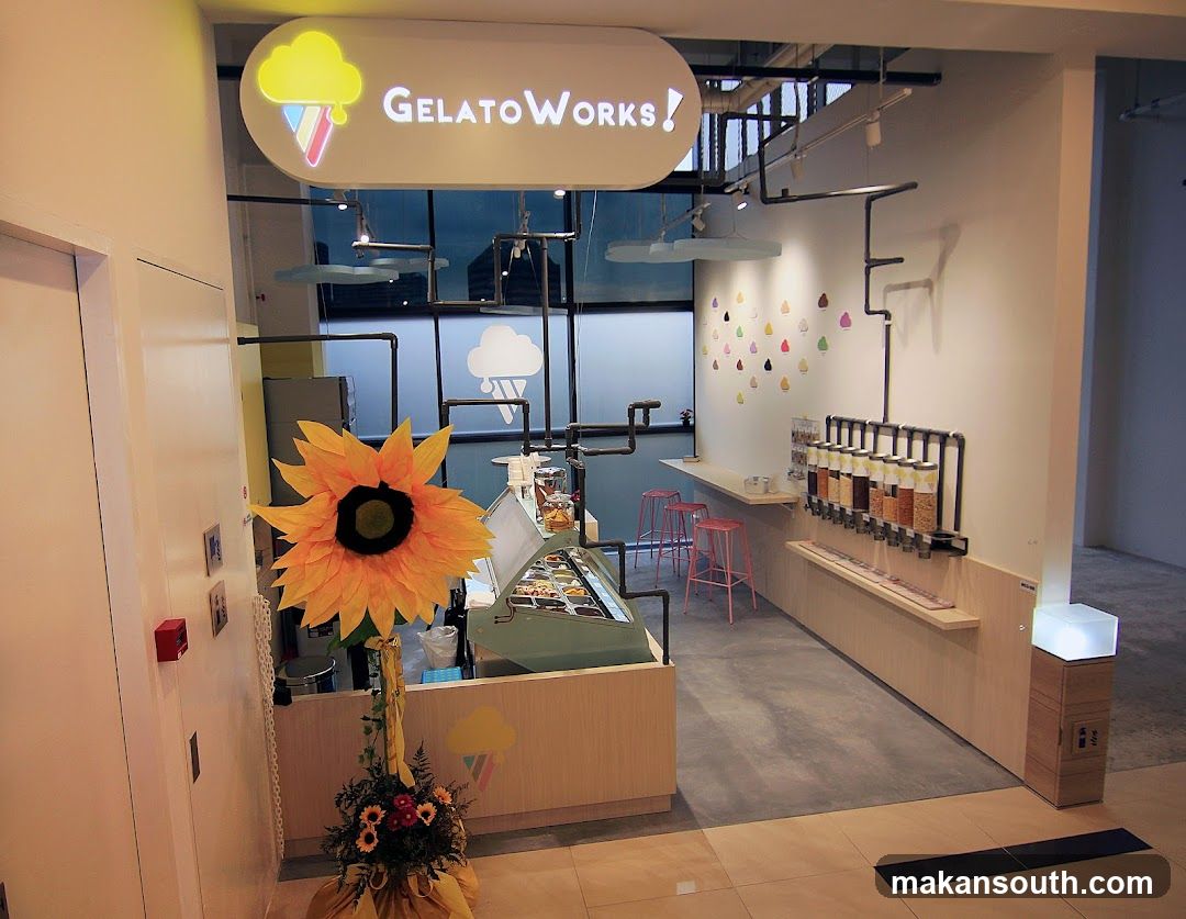 A bright and modern gelato shop interior with a prominent "GelatoWorks!" sign above the counter. The shop features a display case filled with gelato, a topping dispenser station, and pink bar stools. A large artificial sunflower and a floral arrangement are placed near the entrance. The overall ambiance is clean and inviting.