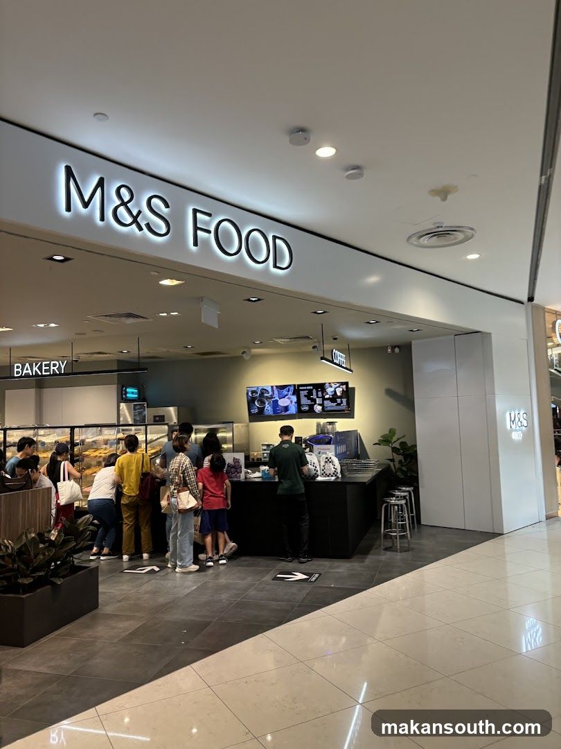 A view of the M&S Food storefront inside a mall. The prominent sign "M&S FOOD" is illuminated above the entrance. To the left, a "BAKERY" sign is visible, with a display case filled with baked goods. A counter area with staff and customers is in the center, and a "COFFEE" sign hangs above. To the right, another "M&S FOOD" logo is displayed on a pillar.