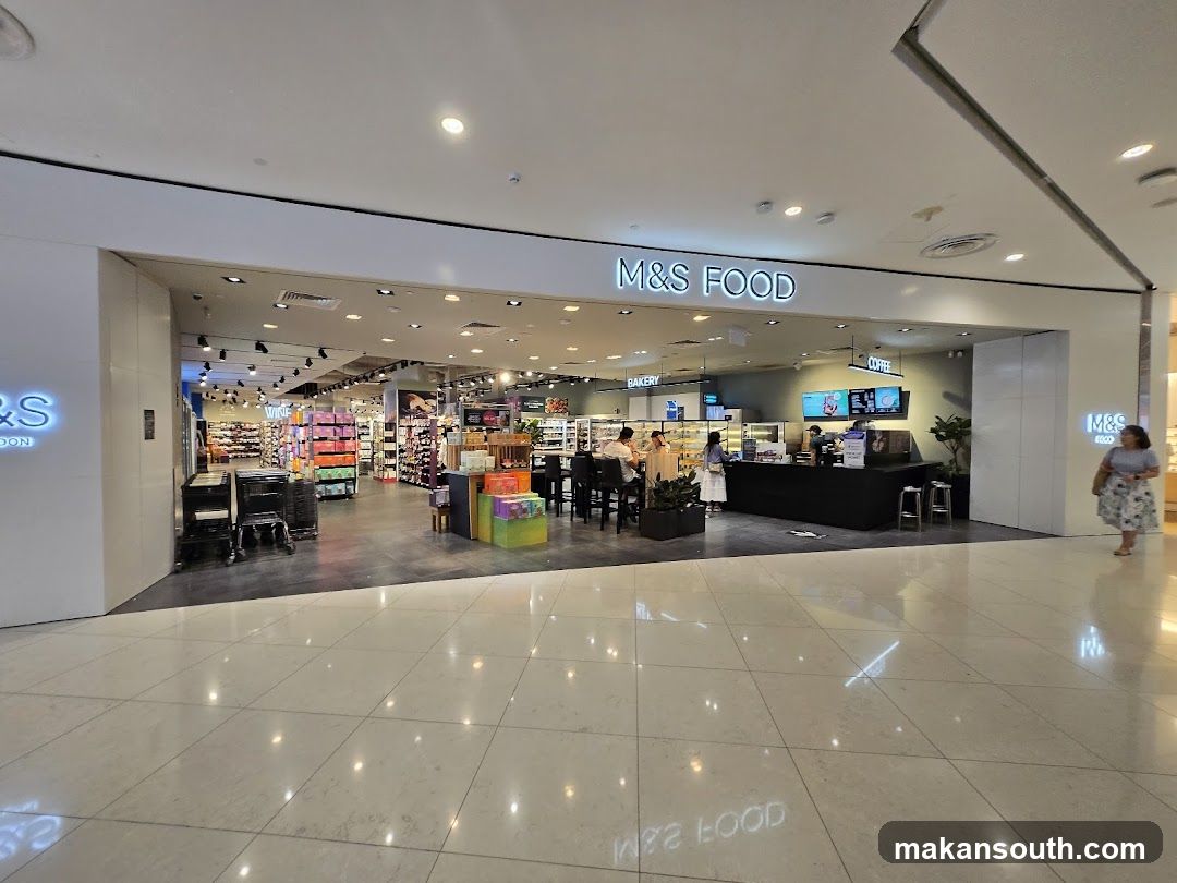 The exterior of an M&S Food store in a shopping mall. The store has a large open entrance with signage above that reads "M&S FOOD". Inside, shelves are stocked with various food items, and there is a coffee counter and bakery section. Several customers are visible inside the store.