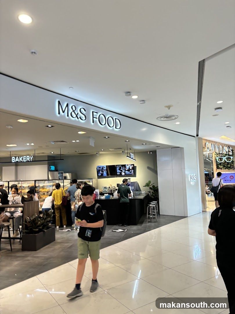The exterior of an M&S FOOD shop with a prominent sign. To the left, a sign reads 'BAKERY'. People are visible inside the shop, browsing and ordering.