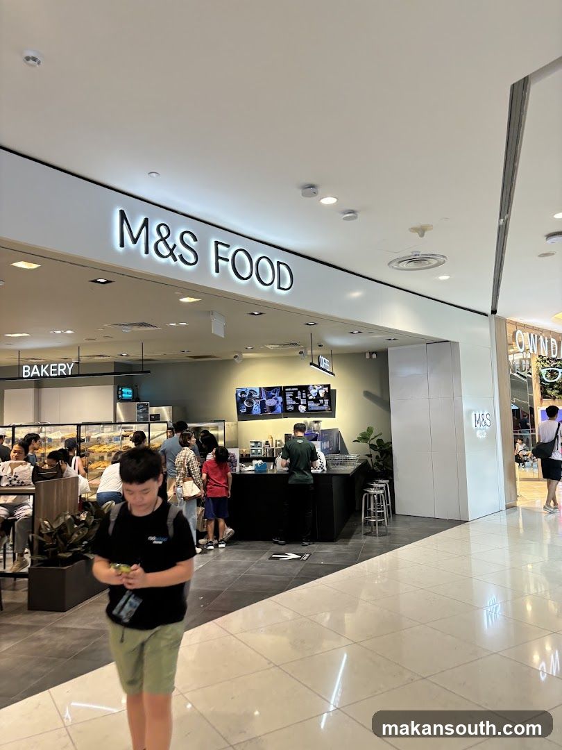 A view of the M&S FOOD storefront in a mall. The signage "M&S FOOD" is prominently displayed above the entrance. To the left, a "BAKERY" sign is visible, and to the right, a section of the store with "M&S" branding is seen. Inside, a coffee counter with customers and staff is visible, along with display cases of baked goods.