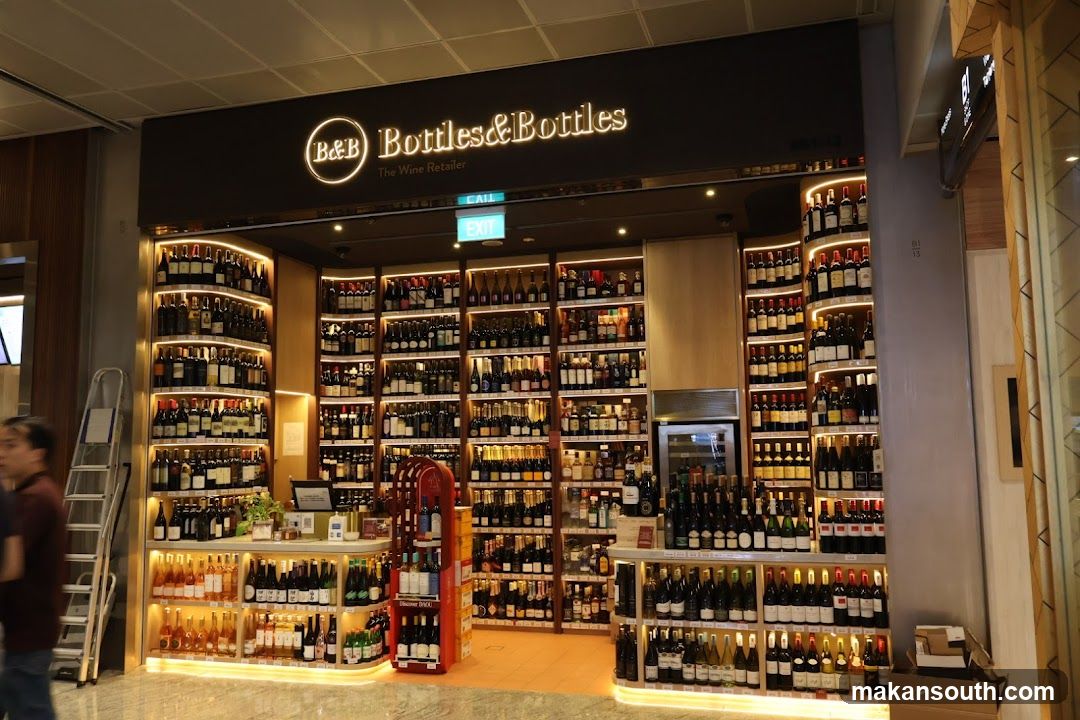A well-lit wine shop with shelves stocked with numerous bottles of wine. The shop's name, "B&B Bottles & Bottles The Wine Retailer," is prominently displayed above the entrance. Various types of wine bottles are arranged on shelves, with some displayed on tiered stands at the front. An exit sign is visible above the entrance.