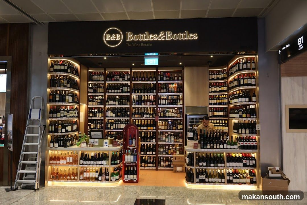The exterior of a wine retail store called "Bottles & Bottles The Wine Retailer". The store is well-lit and features extensive shelving filled with numerous bottles of wine. A staff member is visible behind the counter.