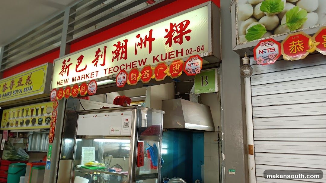 A storefront with a large illuminated sign that reads "NEW MARKET TEOCHEW KUEH" in English and Chinese characters. Below the main sign, there are decorative hexagonal banners with Chinese characters and the NETS logo. To the left, another sign advertises "BAHRU SOYA BEAN" with images of drinks. The shop appears to be a food stall with kitchen equipment visible inside.