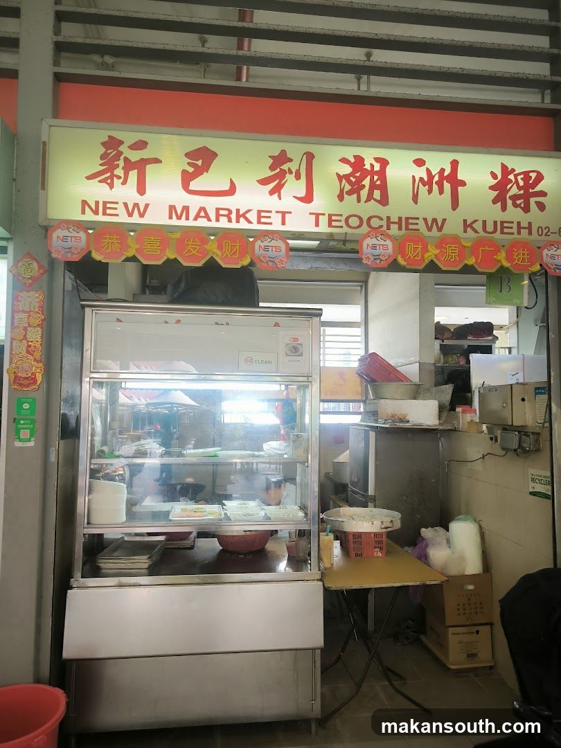 A shop front with a sign that reads "NEW MARKET TEOCHEW KUEH" in English and Chinese characters. Below the main sign, there are decorative banners with Chinese phrases and the NETS logo. A glass display case filled with food items is visible, along with a small table and various boxes and containers.