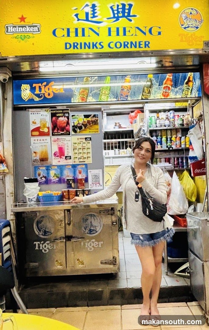A woman stands in front of a brightly lit drinks stall. The stall has a yellow awning with the words "CHIN HENG DRINKS CORNER" and logos for Heineken and Tiger beer. Below the awning, bottles of beer are displayed. A cooler with "Tiger" branding is in the foreground. Various drinks and posters are visible on the stall's wall.