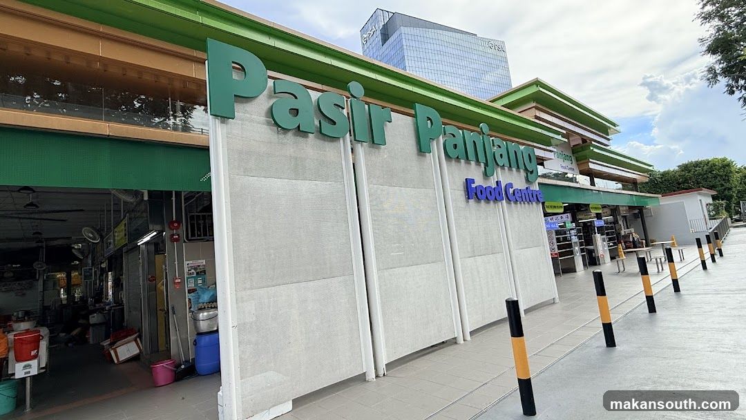 The exterior of the Pasir Panjang Food Centre with its prominent green signage. The building has a modern design with a green roof and beige facade. Several stalls are visible inside the open-air food centre, with tables and chairs set up outside. A tall glass building is visible in the background.