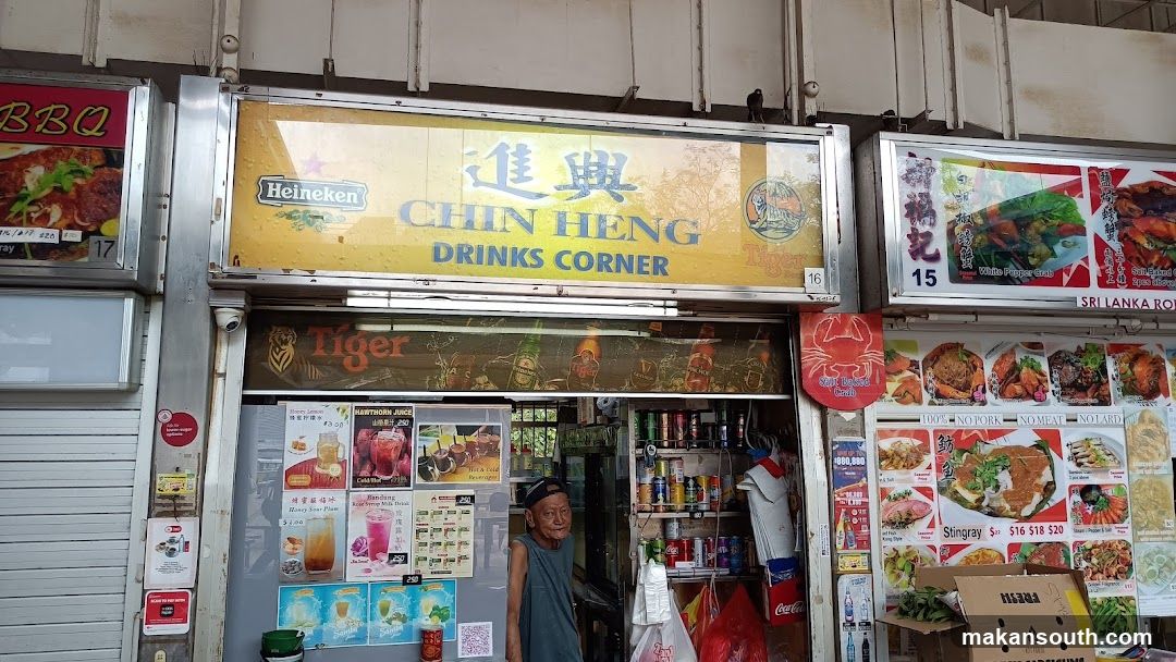 A storefront for Chin Heng Drinks Corner, featuring a large yellow sign with the business name and logos for Heineken and Tiger beer. Below the main sign is a display of various drinks and food items, including pictures of BBQ, crab, and other dishes. A man stands in the doorway. Shelves inside are stocked with canned drinks.