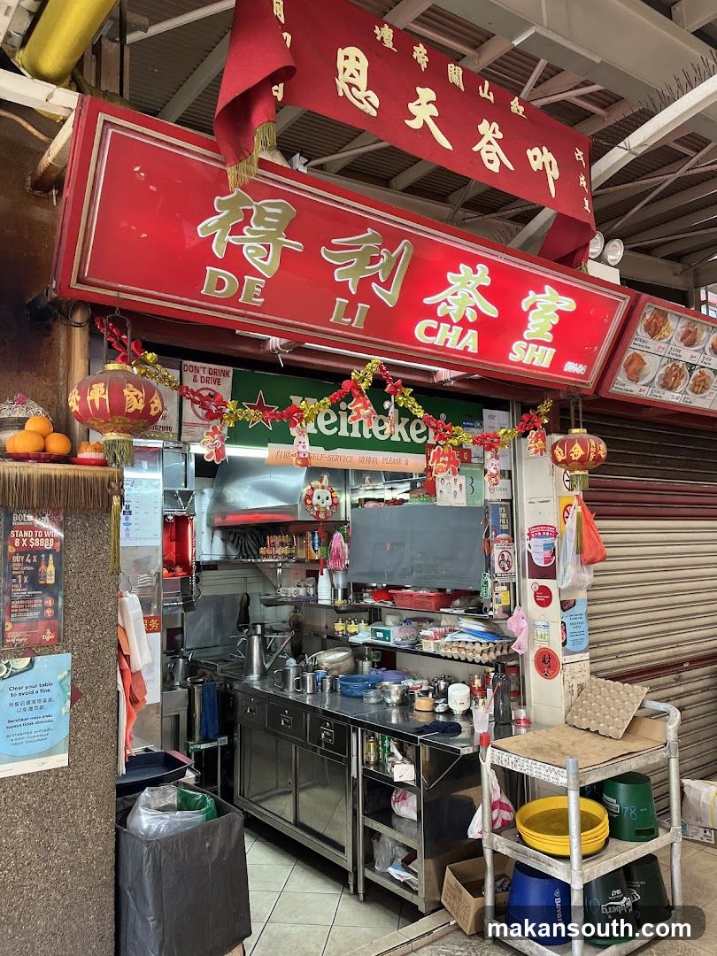 A food stall with a red sign that says "De Li Cha Shi" in gold lettering. There are Chinese characters above the sign. The stall is decorated with red lanterns and festive garlands. The interior of the stall shows a stainless steel counter with various cooking utensils and ingredients. There are also menus and promotional posters visible.