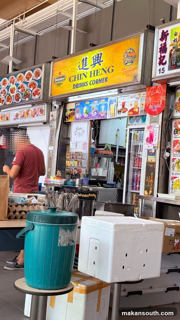 A drinks stall with a large yellow sign that reads "CHIN HENG DRINKS CORNER" and features the Heineken and Tiger logos. Below the sign are various colorful pictures of drinks and food items. A person is visible behind the counter, and a large teal cooler is in the foreground.
