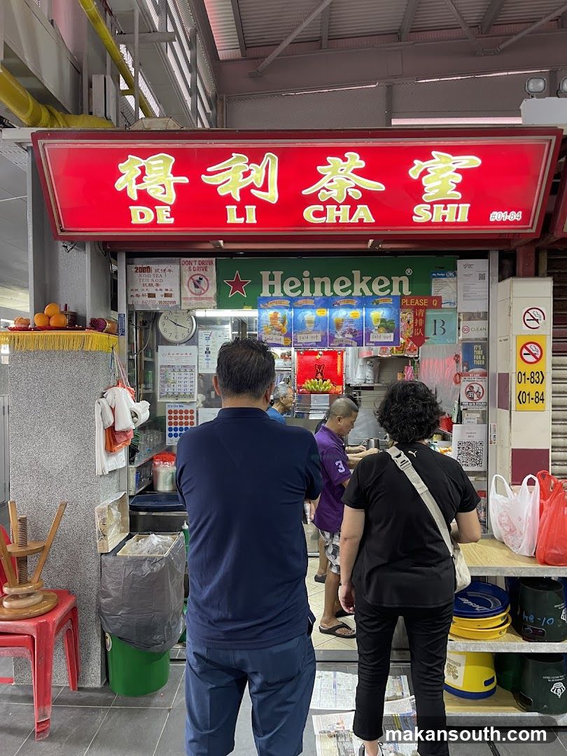A red illuminated sign with Chinese characters and the words "DE LI CHA SHI" is displayed above a food stall. Below the sign, a green Heineken banner is visible. Several people are standing in front of the stall, appearing to order food. The stall has various items displayed, including drinks and what appear to be food items. There are also signs indicating "SOLD OUT" on some of the drink displays.