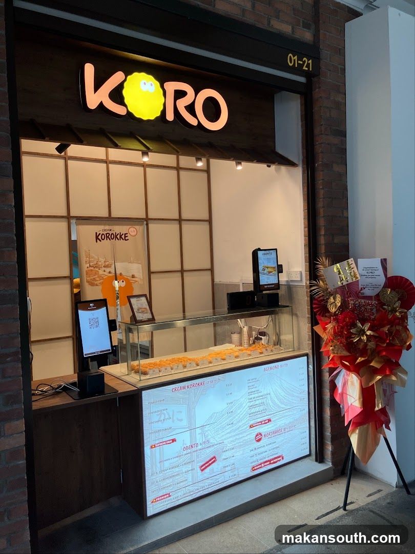 The exterior of a food stall with a prominent illuminated sign that reads "KORO" with a yellow character. Below the sign, a display case shows rows of golden-brown fried food items. A digital menu board is integrated into the front of the counter, displaying various food categories and prices. To the right, a large floral arrangement in red and gold tones is placed near the entrance.