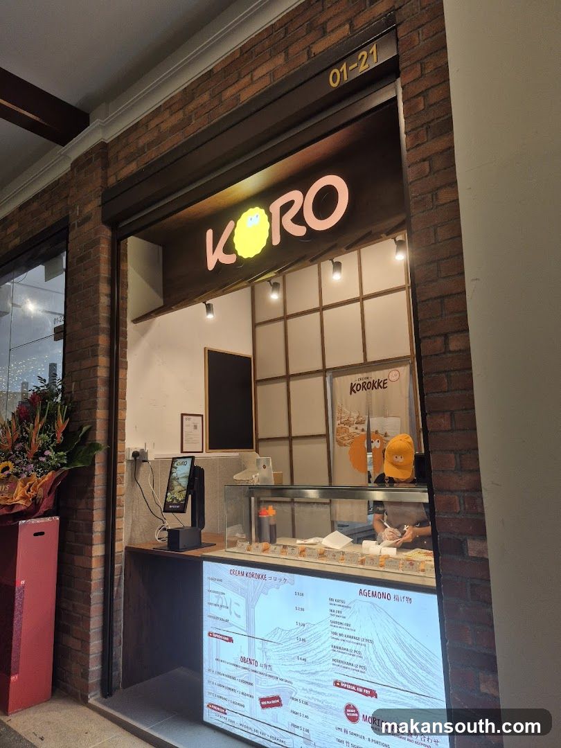 The storefront of a shop called Koro, with a large illuminated sign displaying the name and a cartoon character. The shop has a brick facade and a display window showing food items. A digital menu is visible below the display.