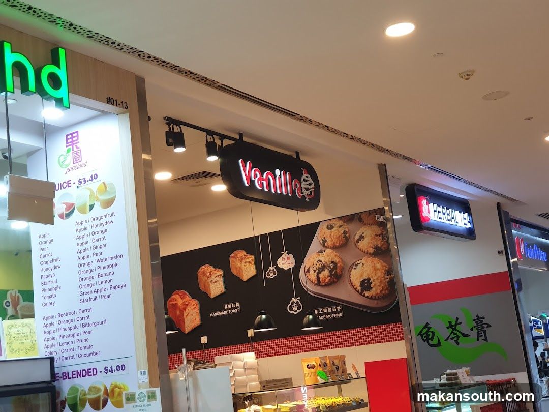 A storefront with two businesses visible. The left side shows a juice bar with a menu board listing various juice combinations. The right side displays a bakery with muffins and toast, and a sign for "Vanilla". Another sign for "Herbal Tea" is also visible.
